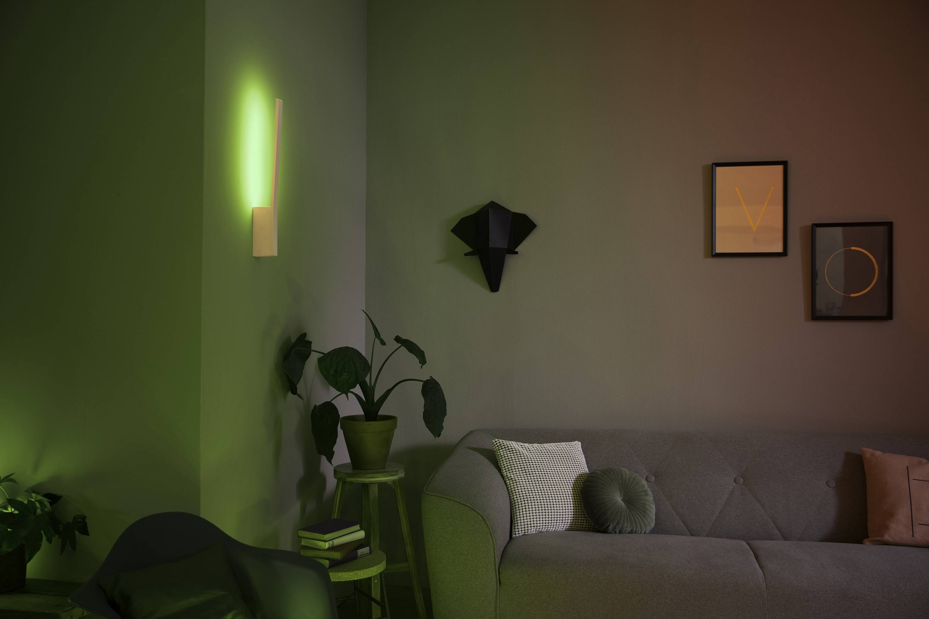 Een woonkamer met een grijze bank, een groene plant, twee wandschilderijen en een groene wandlamp die een sfeervolle verlichting creëert.