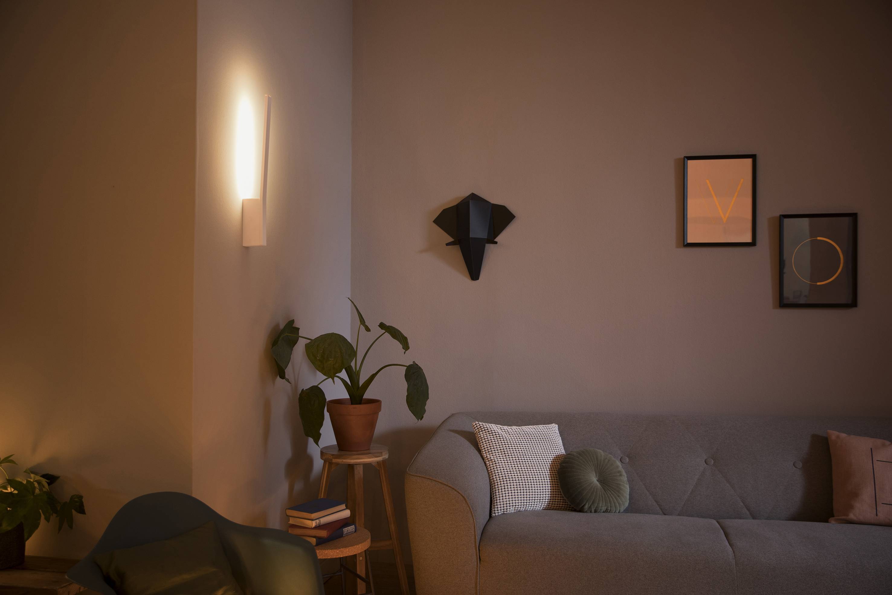 Woonkamer met een grijze bank, een groene fauteuil, een plant op een tafel en moderne wandverlichting; fotolijsten aan de muur.