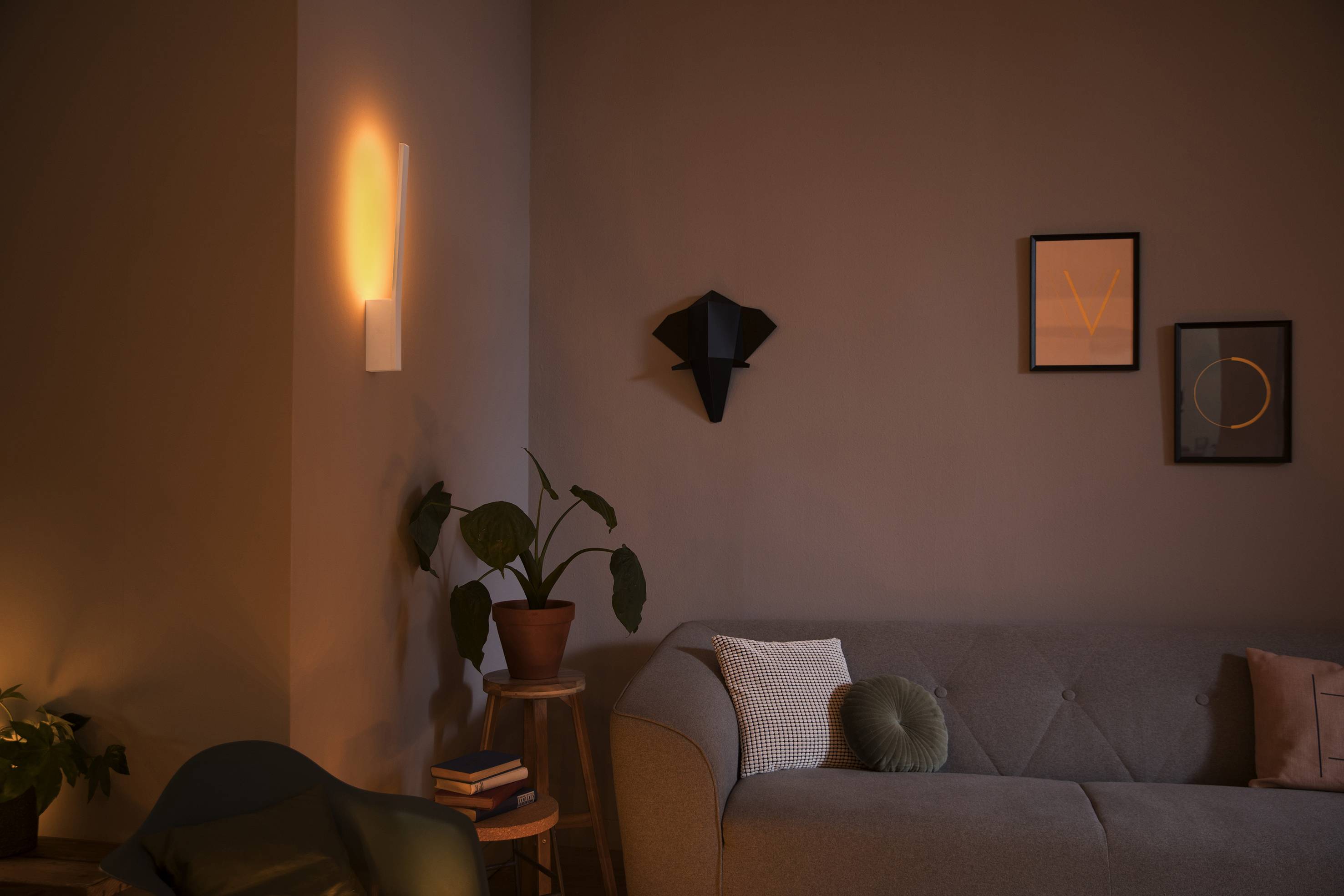 Een woonkamer met gedempt licht. Een wandlamp werpt warm licht op de muur. Een bank met kussens, planten en fotolijsten.