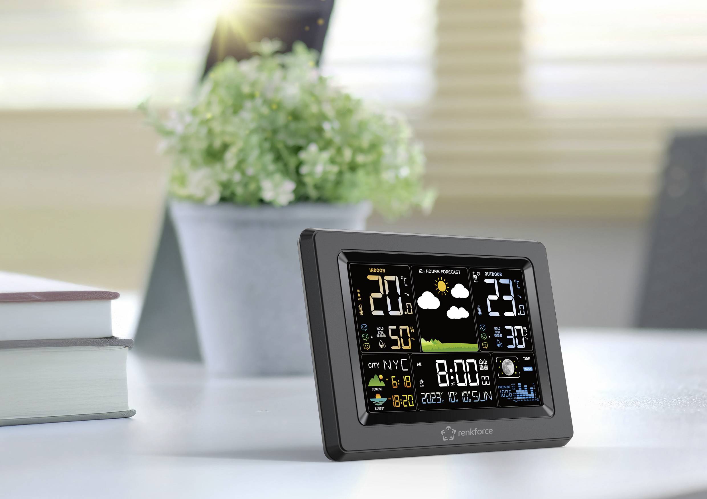 Een digitaal weerstation-scherm toont temperatuur, luchtvochtigheid en weersverwachting, met een plant op de achtergrond.