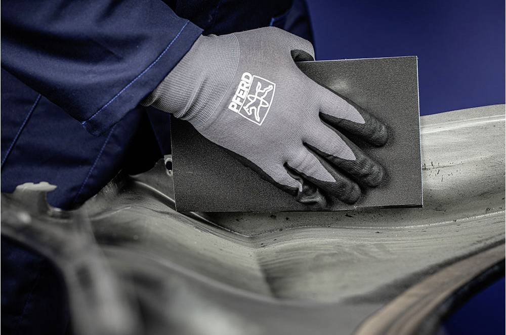 'Arbeider draagt handschoen en slijpt metaaloppervlak met schuurpapier. Beschermende werkkleding en precisie handwerk zijn herkenbaar.'