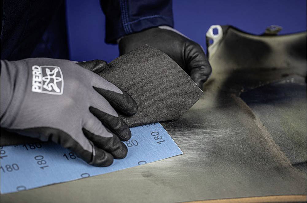 'Twee gehandschoende handen schuren een oppervlak met schuurpapier. Er wordt een blauw schuurpapier met de aanduiding 180 gebruikt.'