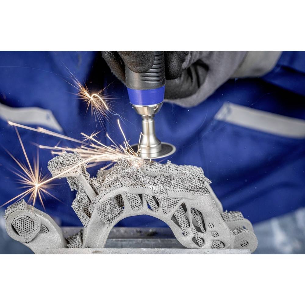 Een persoon slijpt een complex, metallisch 3D-geprint onderdeel terwijl vonken spatten. Ze draagt handschoenen en werkkleding.