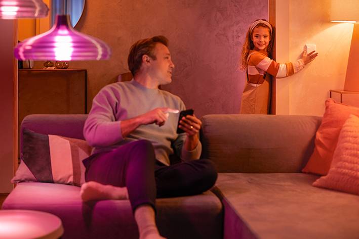 Een man zit op een bank en houdt een smartphone vast, terwijl een lachend meisje op de achtergrond speelt. Woonkamer met roze verlichting.