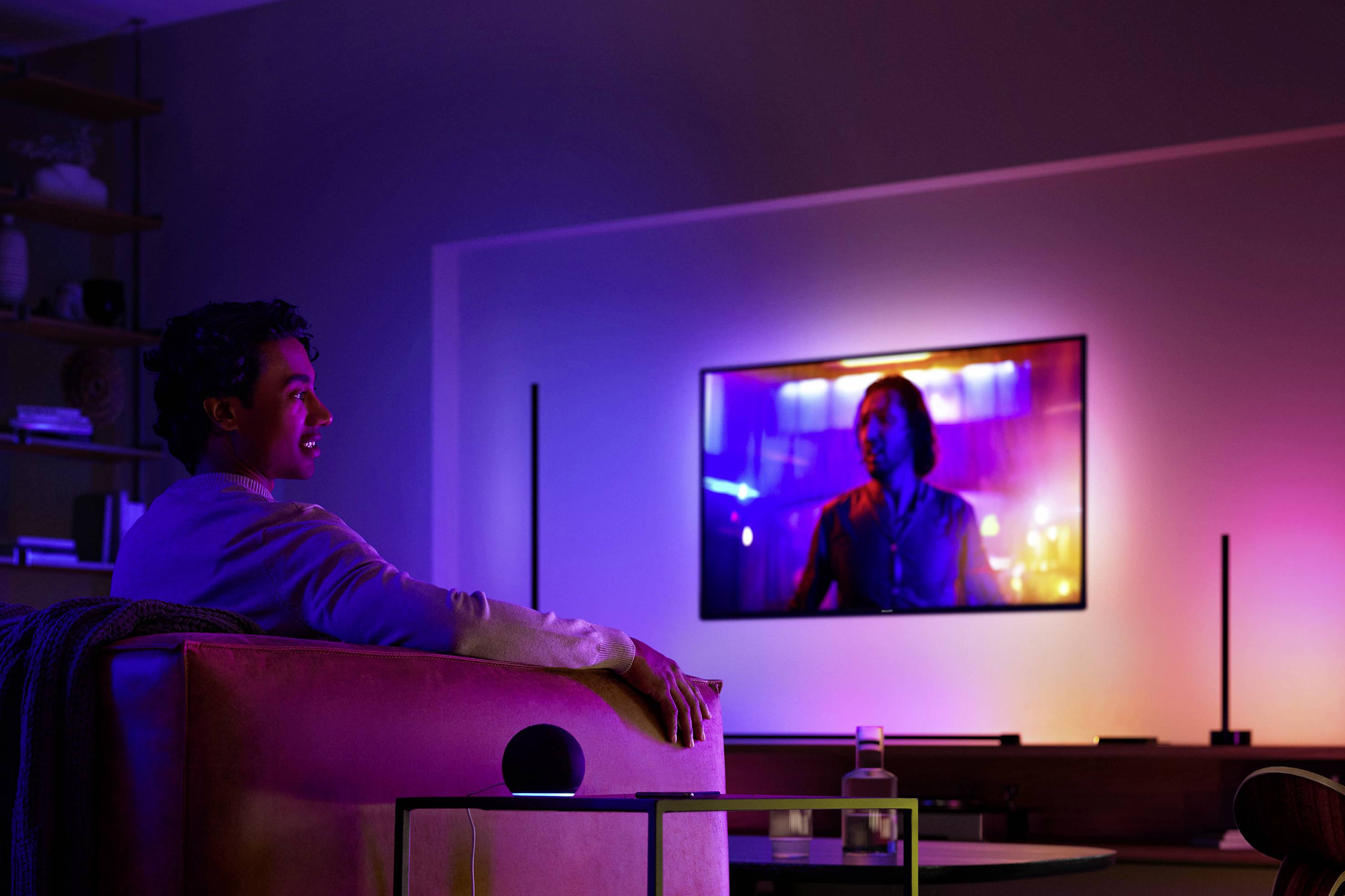 Een persoon zit op een bank en kijkt naar een grote tv aan de muur. De ruimte is verlicht met kleurrijke lichten.