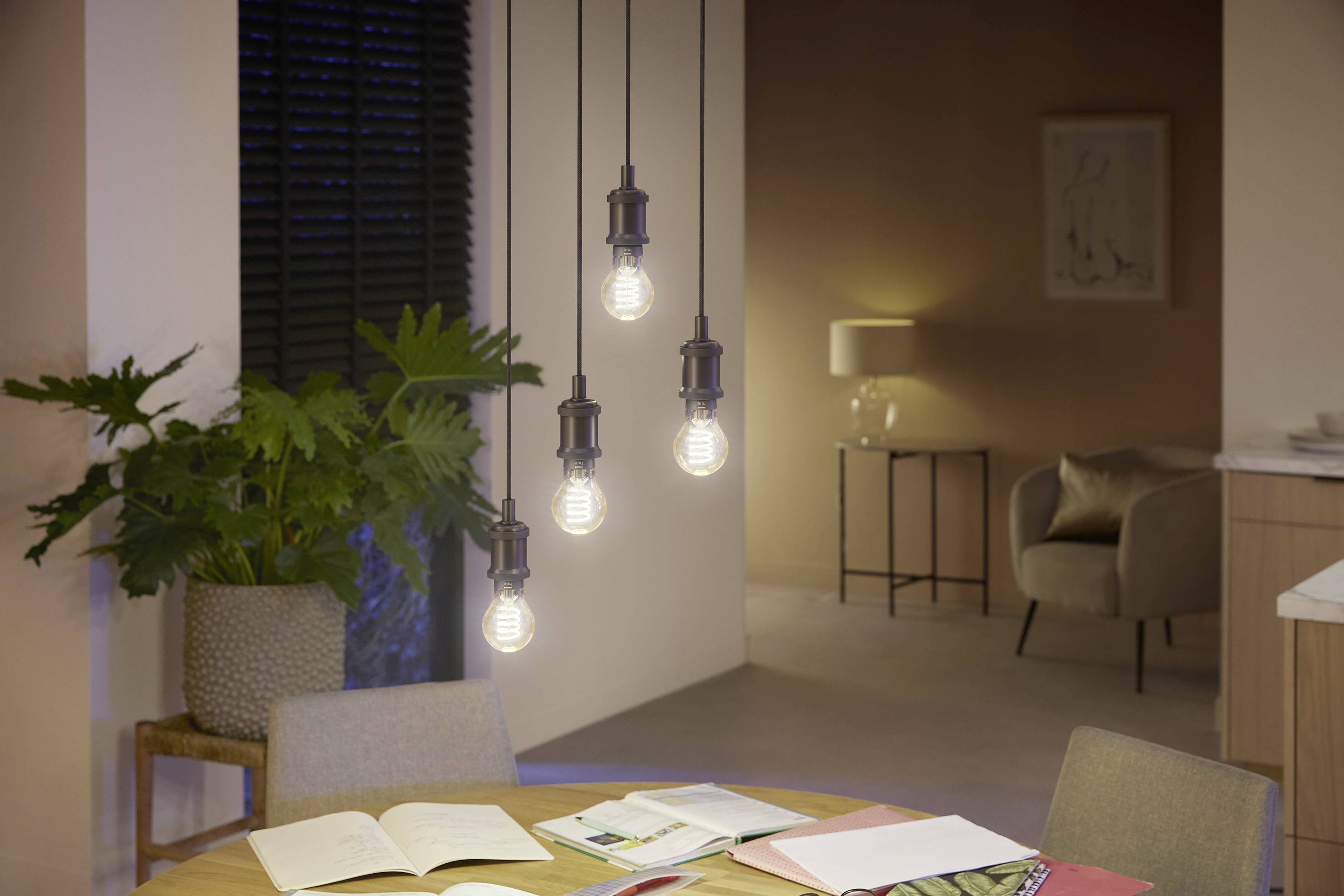 Eetkamer met een ronde tafel en vier hangende lampen boven boeken, op de achtergrond zijn een fauteuil, een staande lamp en een plant te zien.