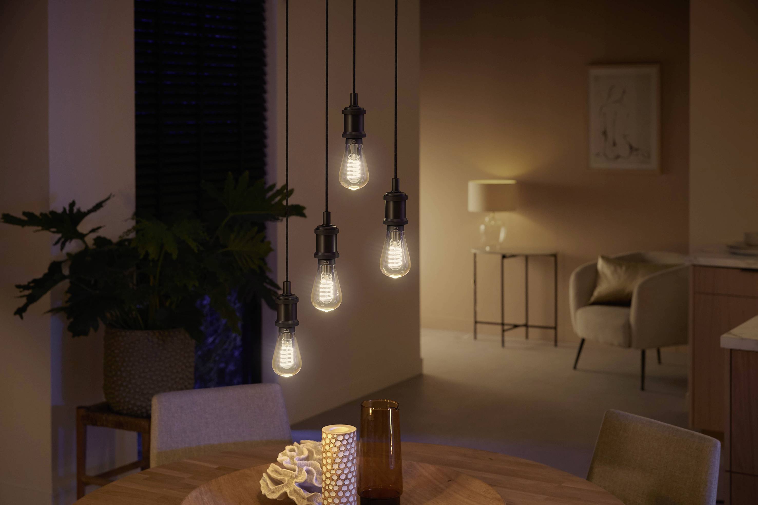 Vijf hangende gloeilampen verlichten een moderne woonruimte met een ronde houten tafel, planten en subtiele verlichting op de achtergrond.