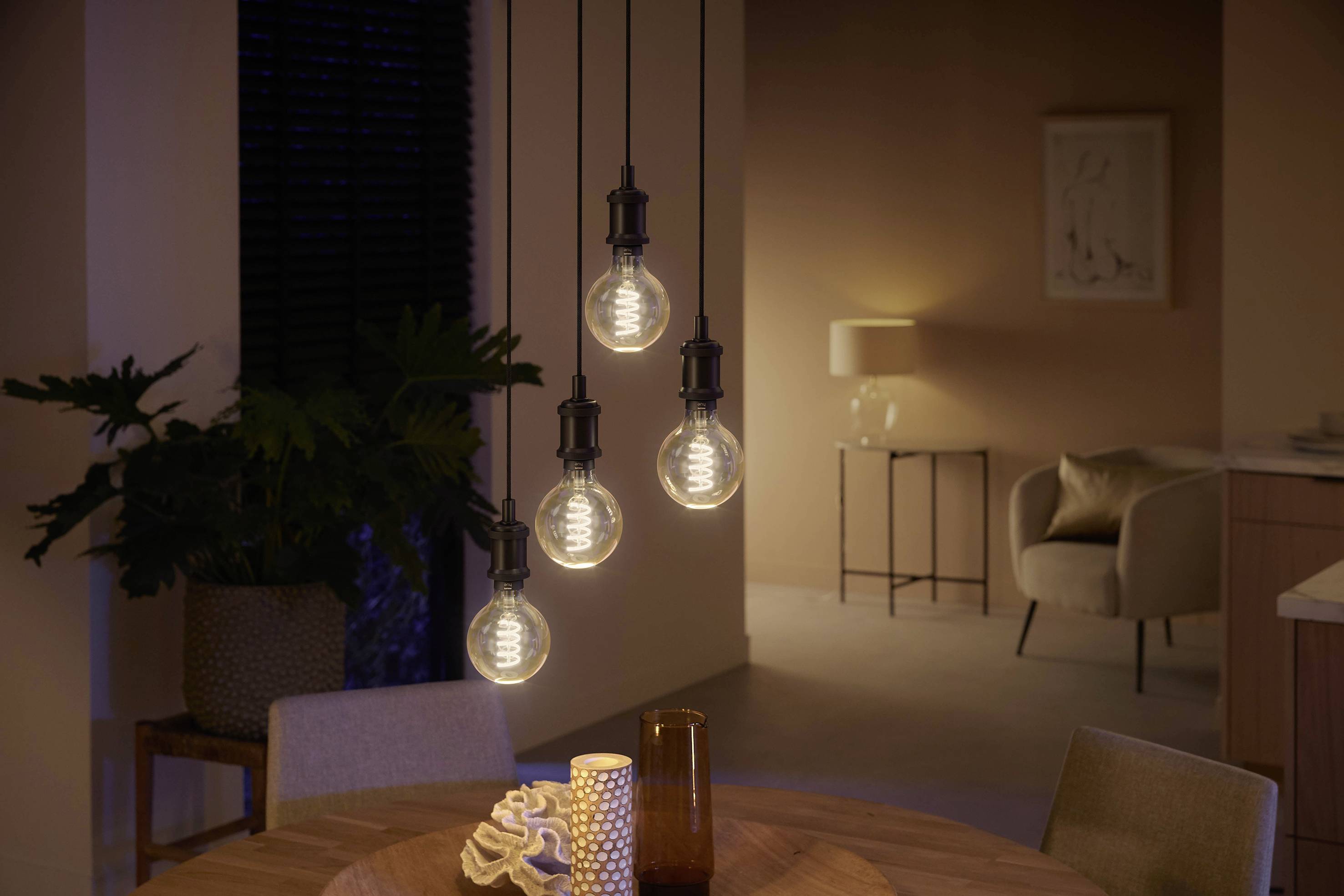 Een gezellig verlichte woonkamer met vier hangende gloeilampen boven een ronde houten tafel. Op de achtergrond zijn planten en meubels.