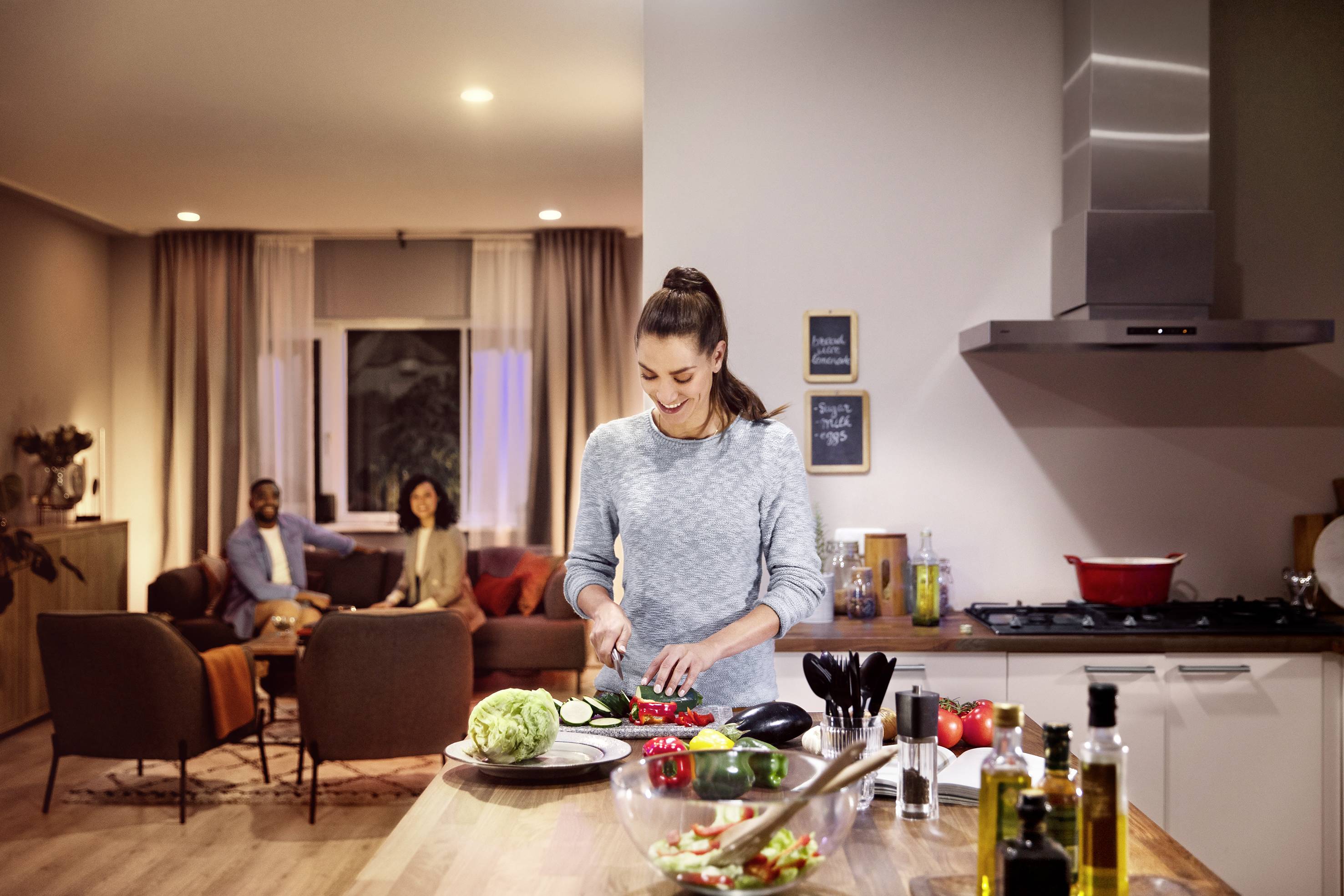 Een vrouw snijdt groenten in een moderne keuken, terwijl op de achtergrond twee personen op een bank zitten te praten.