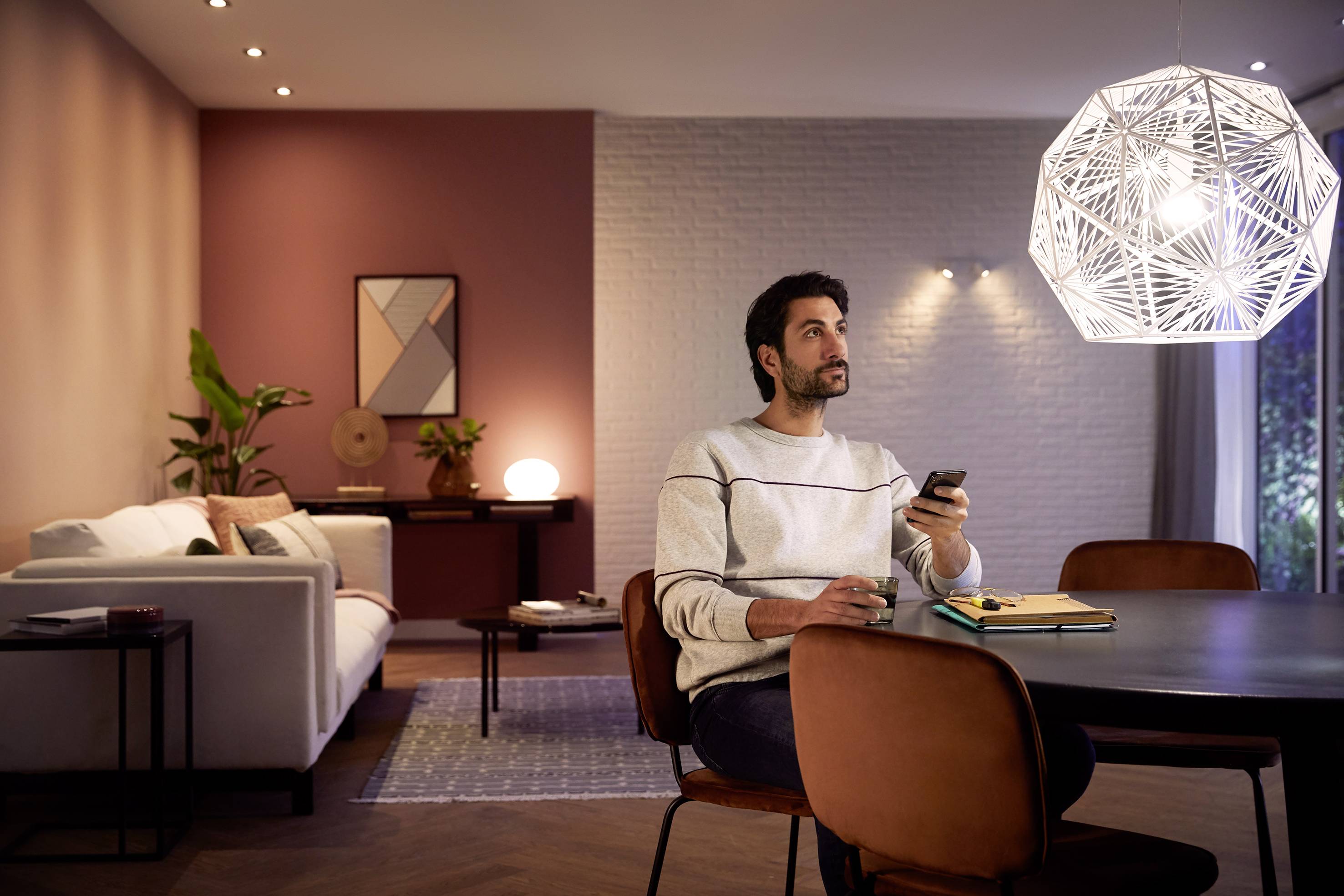 Een man zit nadenkend aan een tafel in een modern, stijlvol ingericht woonkamer. Hij houdt een afstandsbediening vast en kijkt voor zich uit.