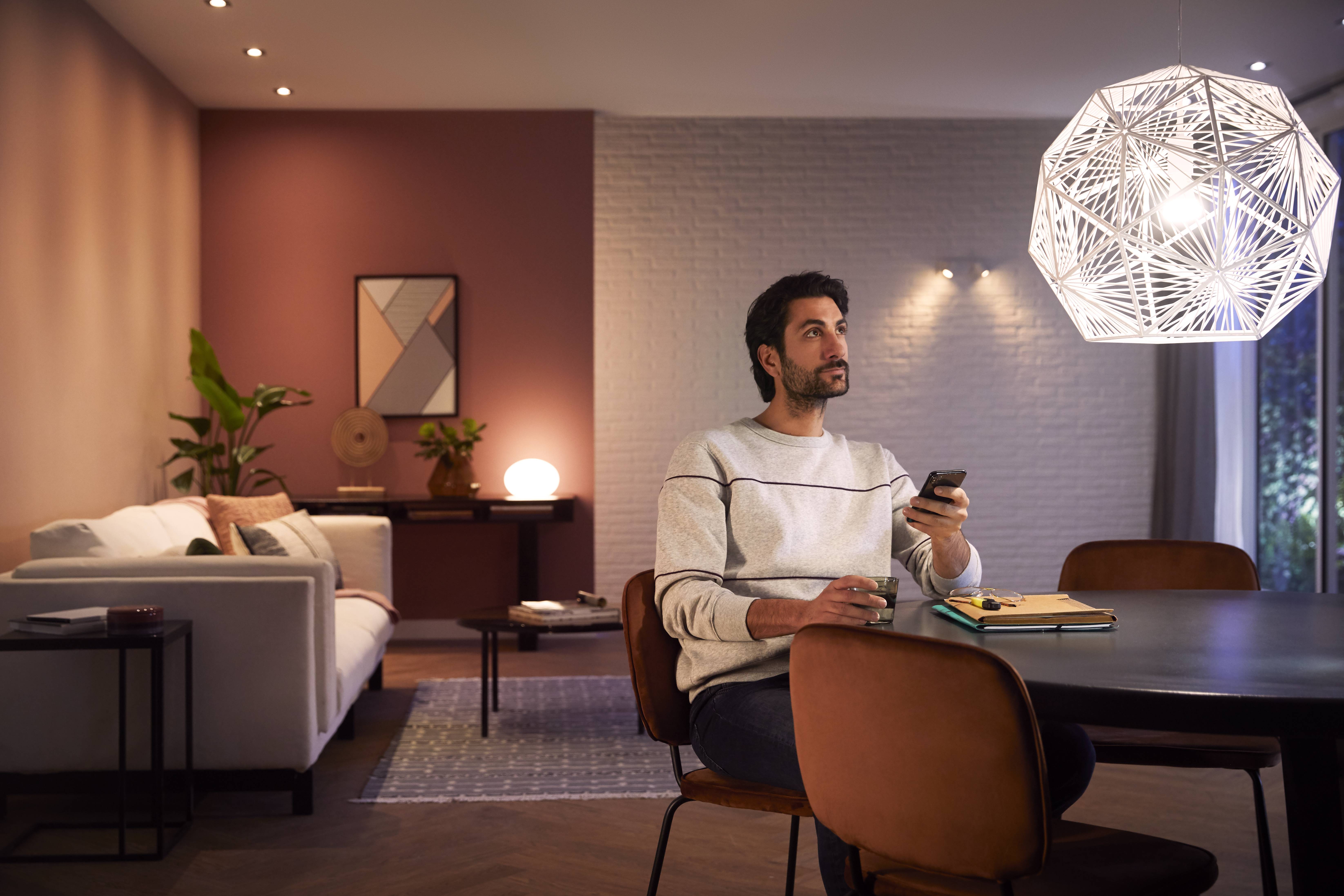 Een man zit aan een tafel in een modern ingericht woonkamer, houdt een afstandsbediening vast en kijkt nadenkend omhoog.