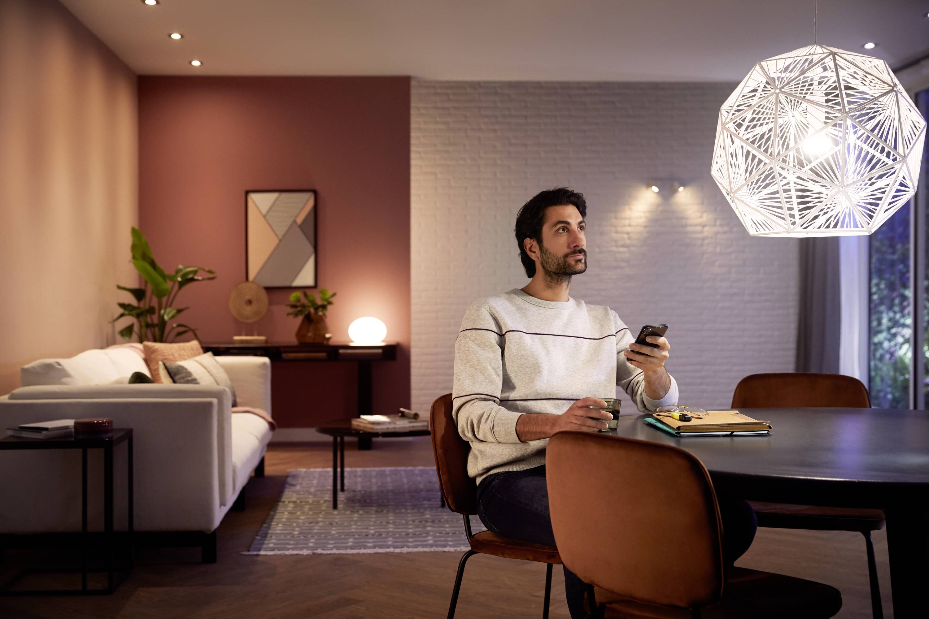 Een man zit aan een tafel met een notitieboek en een pen. Hij houdt een afstandsbediening vast en kijkt nadenkend in een gezellige woonkamer.