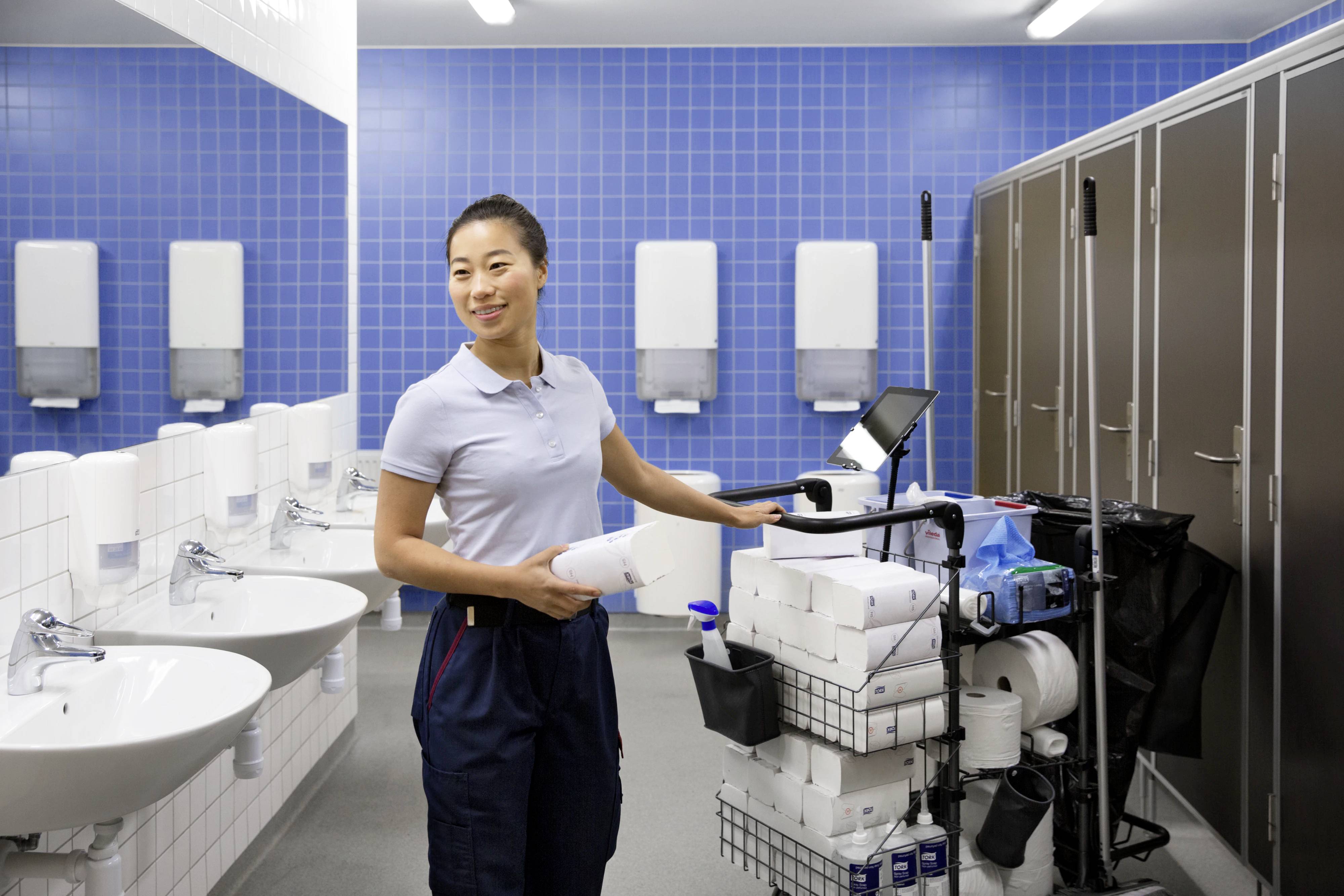 Een schoonmaker staat glimlachend naast een schoonmaakkar in een schoon, betegeld badkamer met meerdere wastafels.