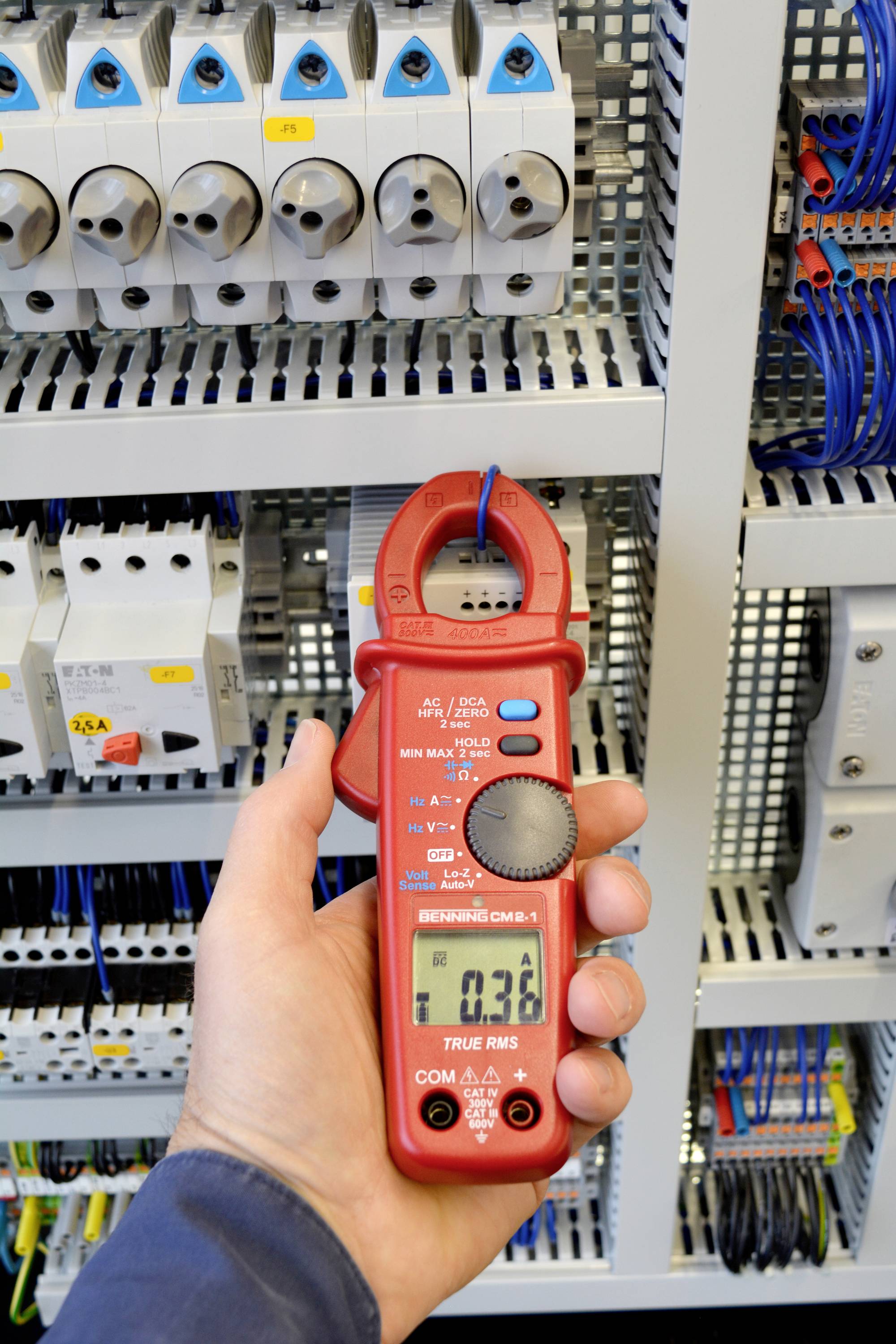 Een persoon meet met een rode digitale multimeter de elektrische stroom in een schakelkast. Het display toont '0,36'.