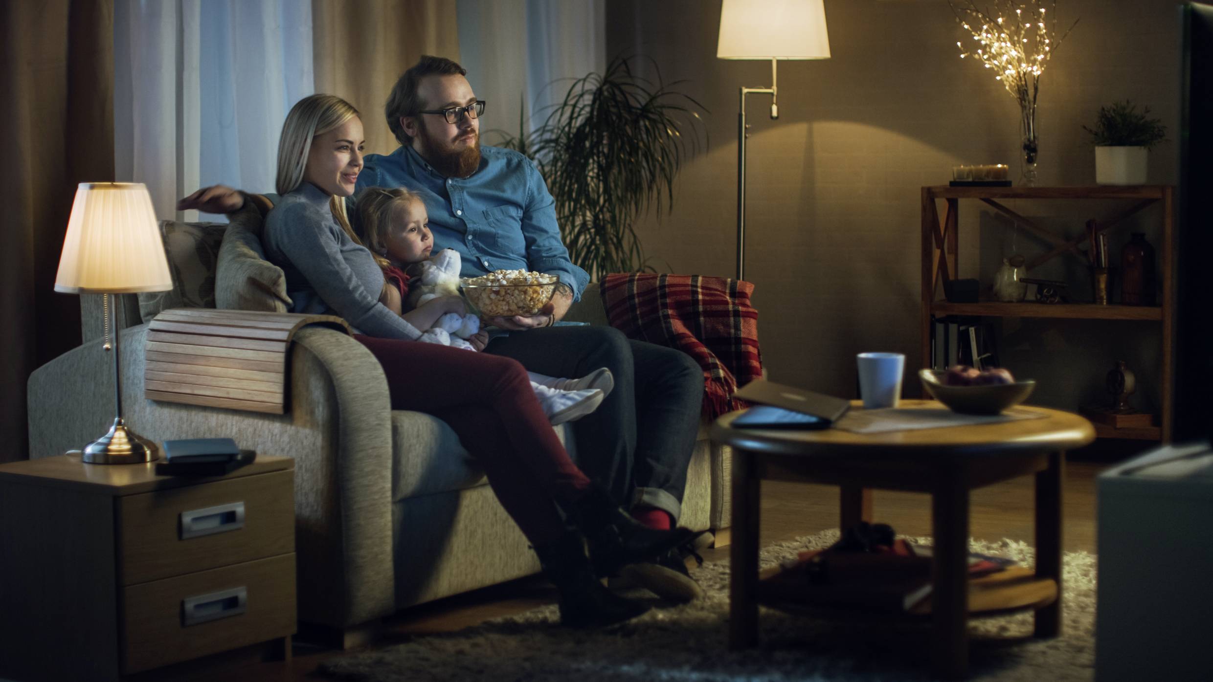 Een familie zit samen op de bank in de woonkamer en kijkt naar een film. Ze zien er ontspannen uit, en op tafel staat een kom popcorn.