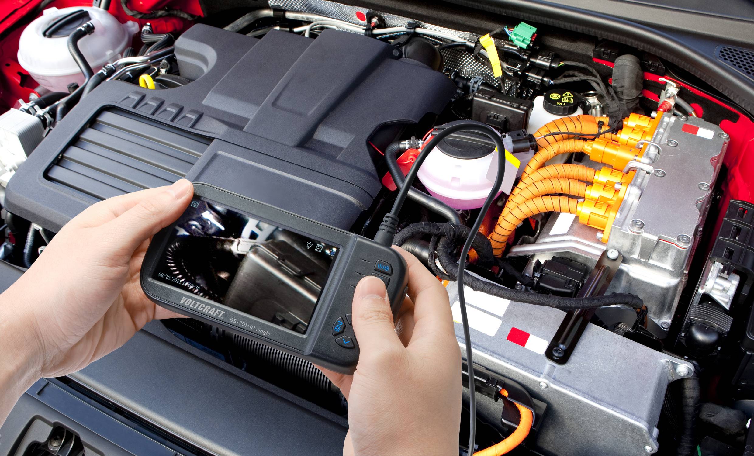 Een persoon houdt een diagnostisch apparaat boven de motor van een auto, waarbij kabels en aansluitingen zichtbaar zijn. Het onderwerp is voertuigonderhoud.