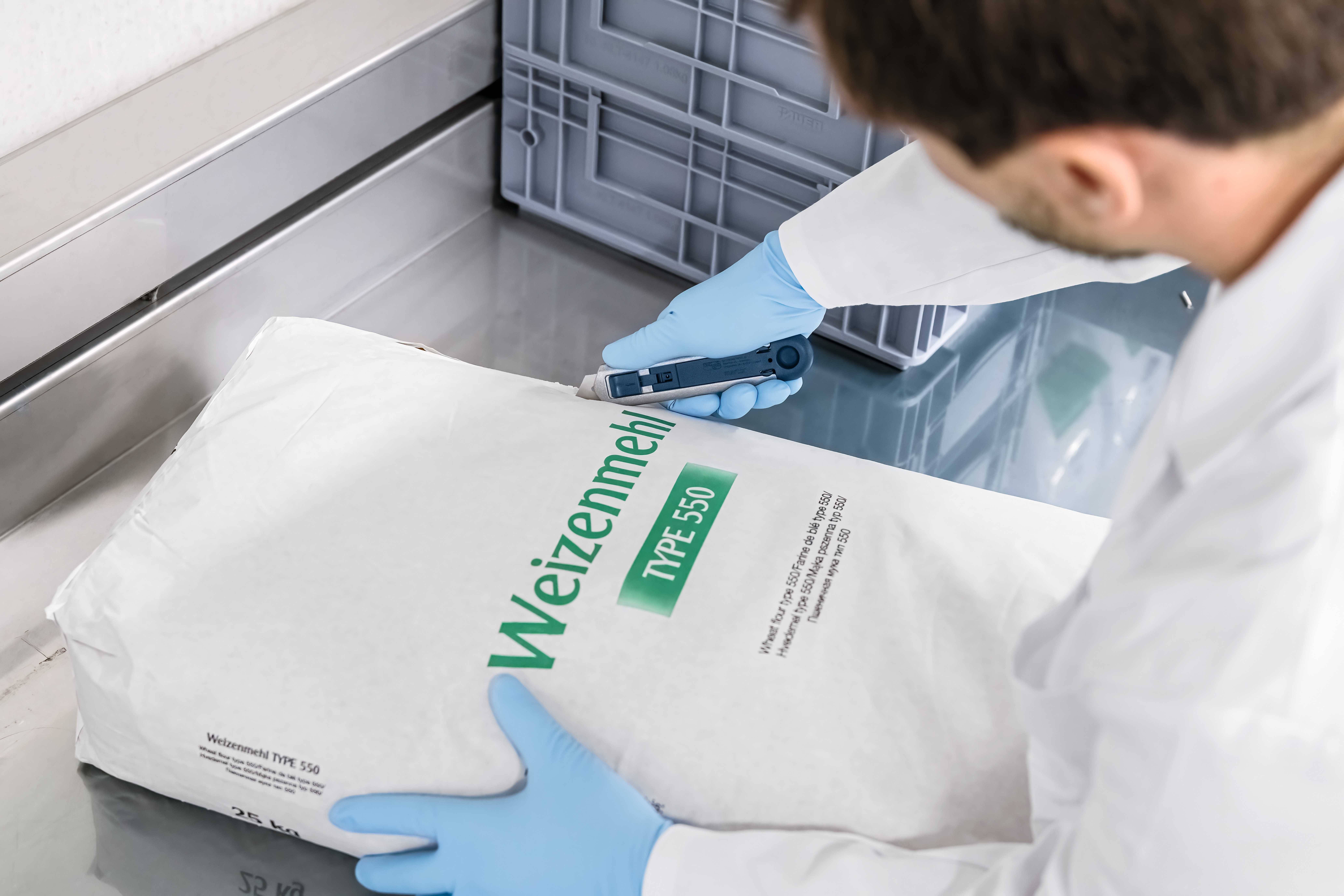 Persoon in witte laborjas controleert een meel verpakking met het opschrift 'Tarwemeel Type 550' op een roestvrijstalen tafel.