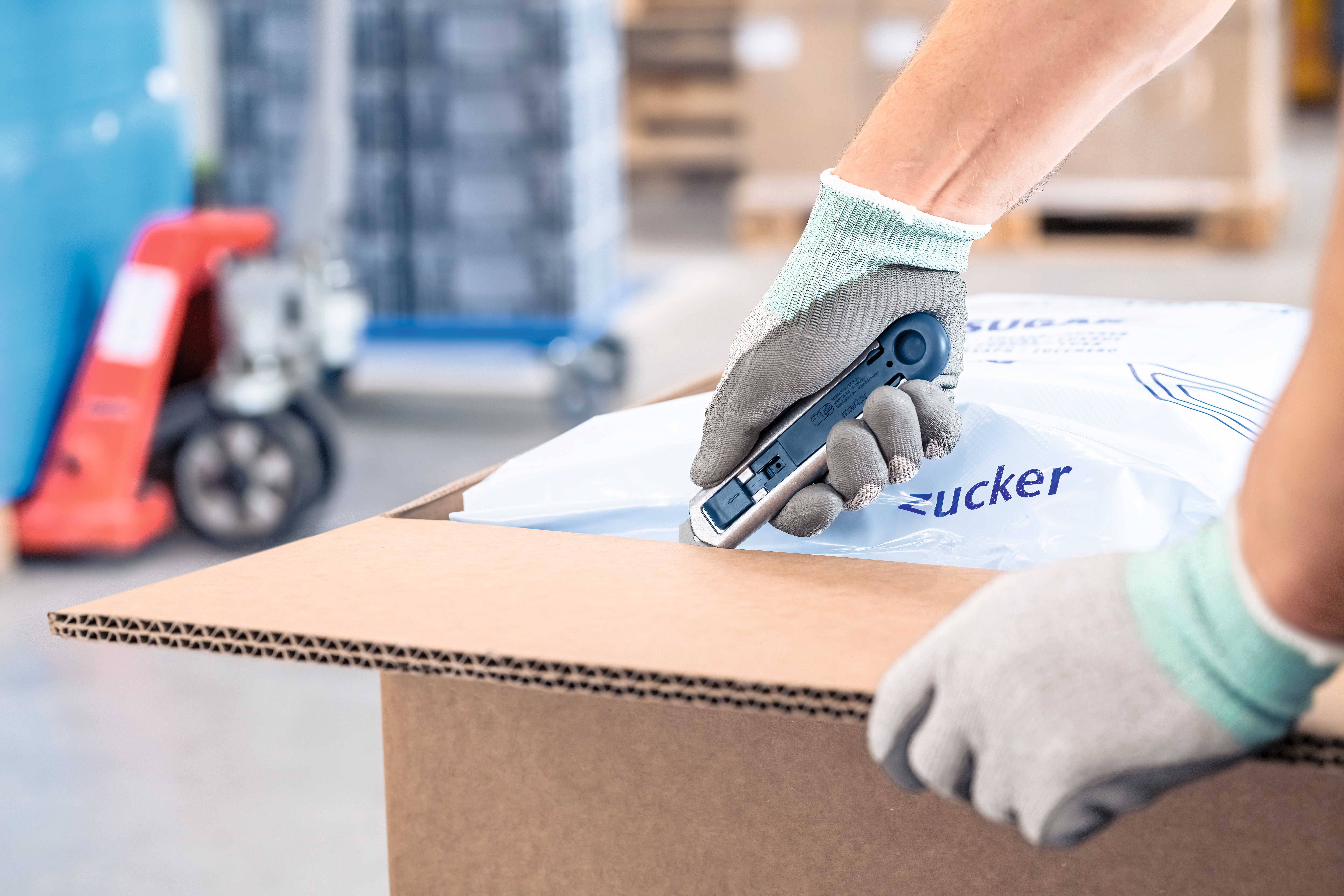 Een persoon snijdt een kartonnen doos open met een veiligheidsmes. De persoon draagt werkhandschoenen. Op de achtergrond staan pallets.