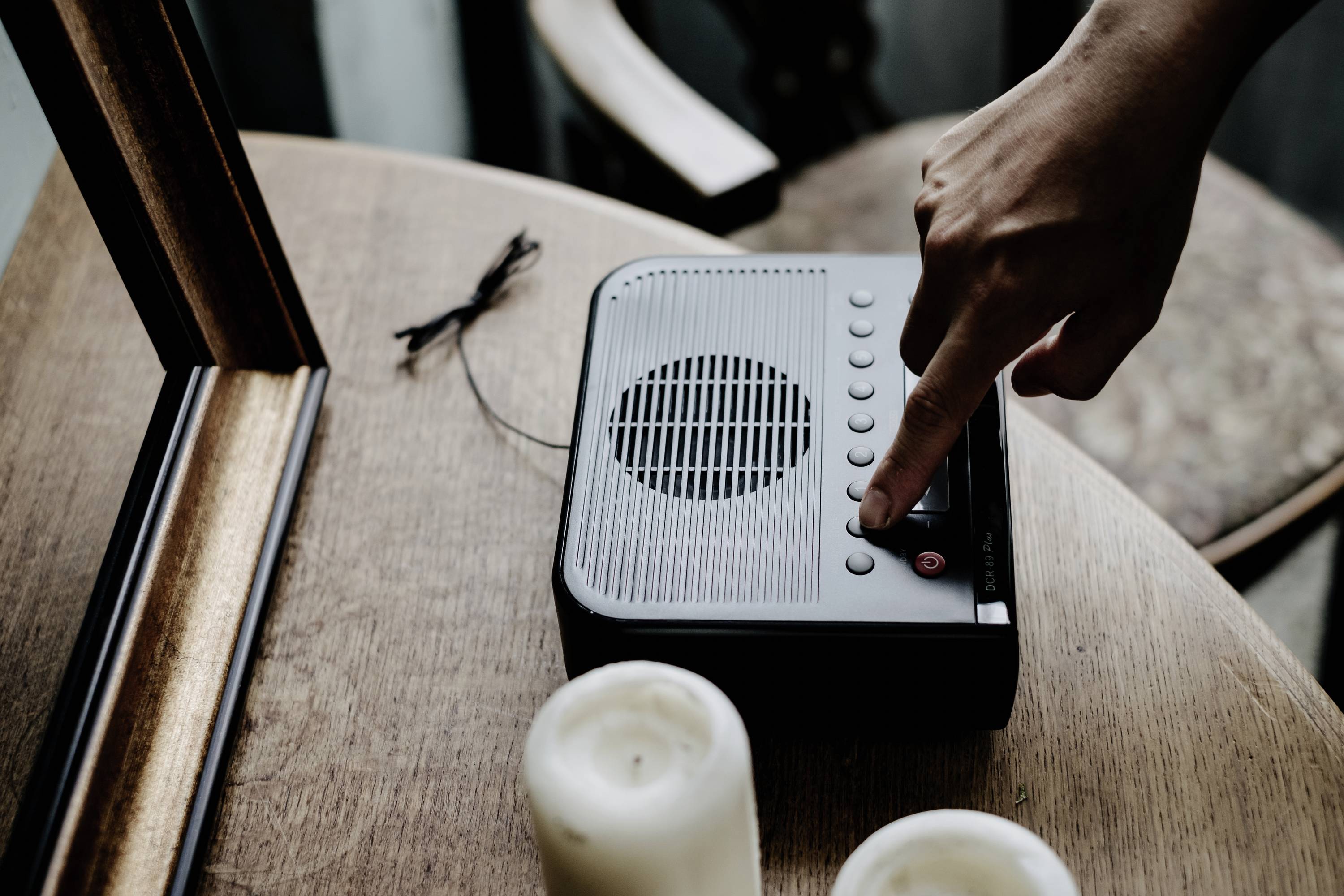 Een hand drukt op een knop van een oude cassetterecorder die op een houten tafel naast twee kaarsen staat.