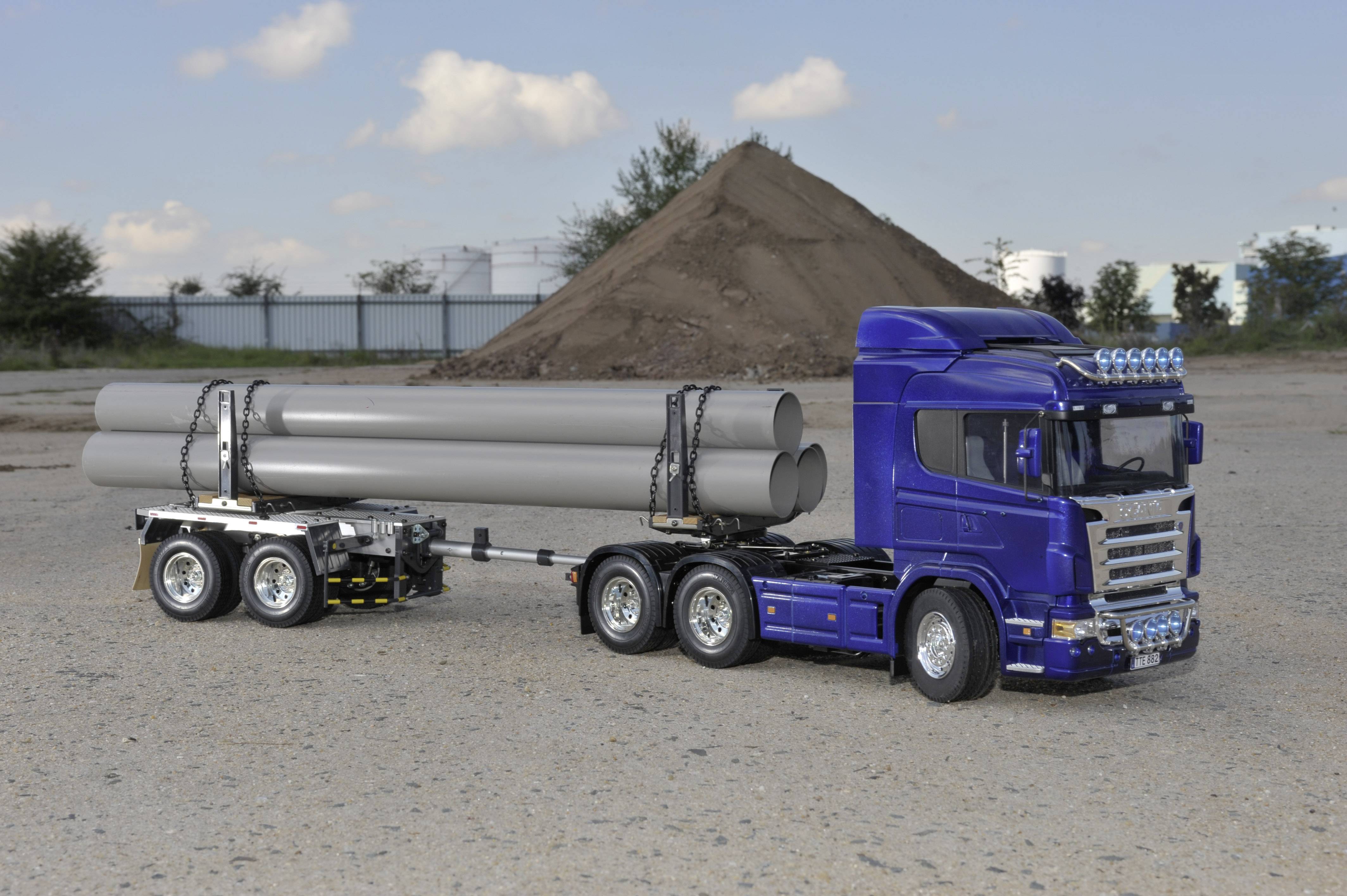 Blauwe vrachtwagen vervoert grote buizen op een aanhanger op een industrieterrein met zandheuvels op de achtergrond.