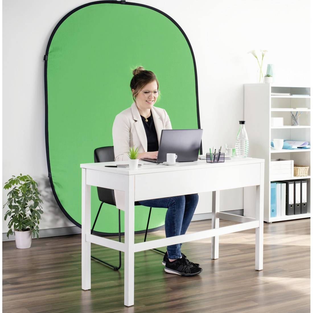 Een vrouw zit aan een witte tafel met een laptop. Achter haar een groene stoffen achtergrond. In de ruimte staan planten en boekenkasten.