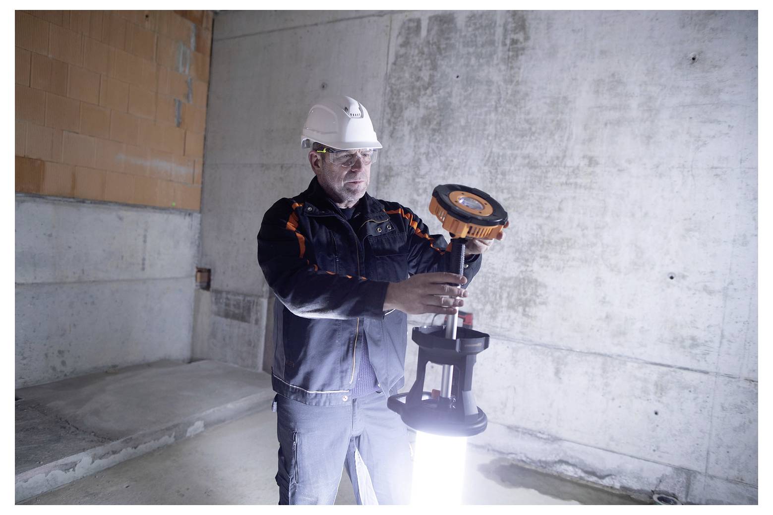 Bouwvakker met een veiligheidshelm stelt een draagbare werklamp bij in een onafgemaakte betonnen kamer.