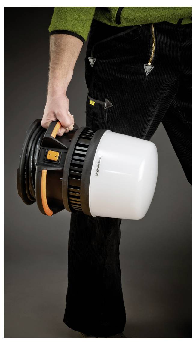 Persoon die een draagbare werklamp bij het handvat vasthoudt, getoond van heup tot knie tegen een donkere achtergrond.