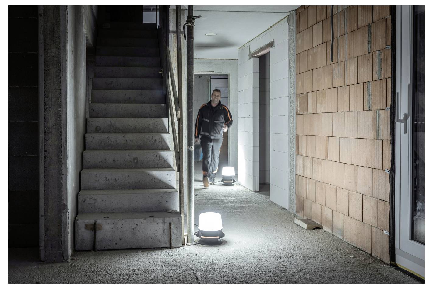 Een werknemer staat in een donkere onafgewerkte gang naast betonnen trappen en verlichte vloerlampen in een gebouw in aanbouw.