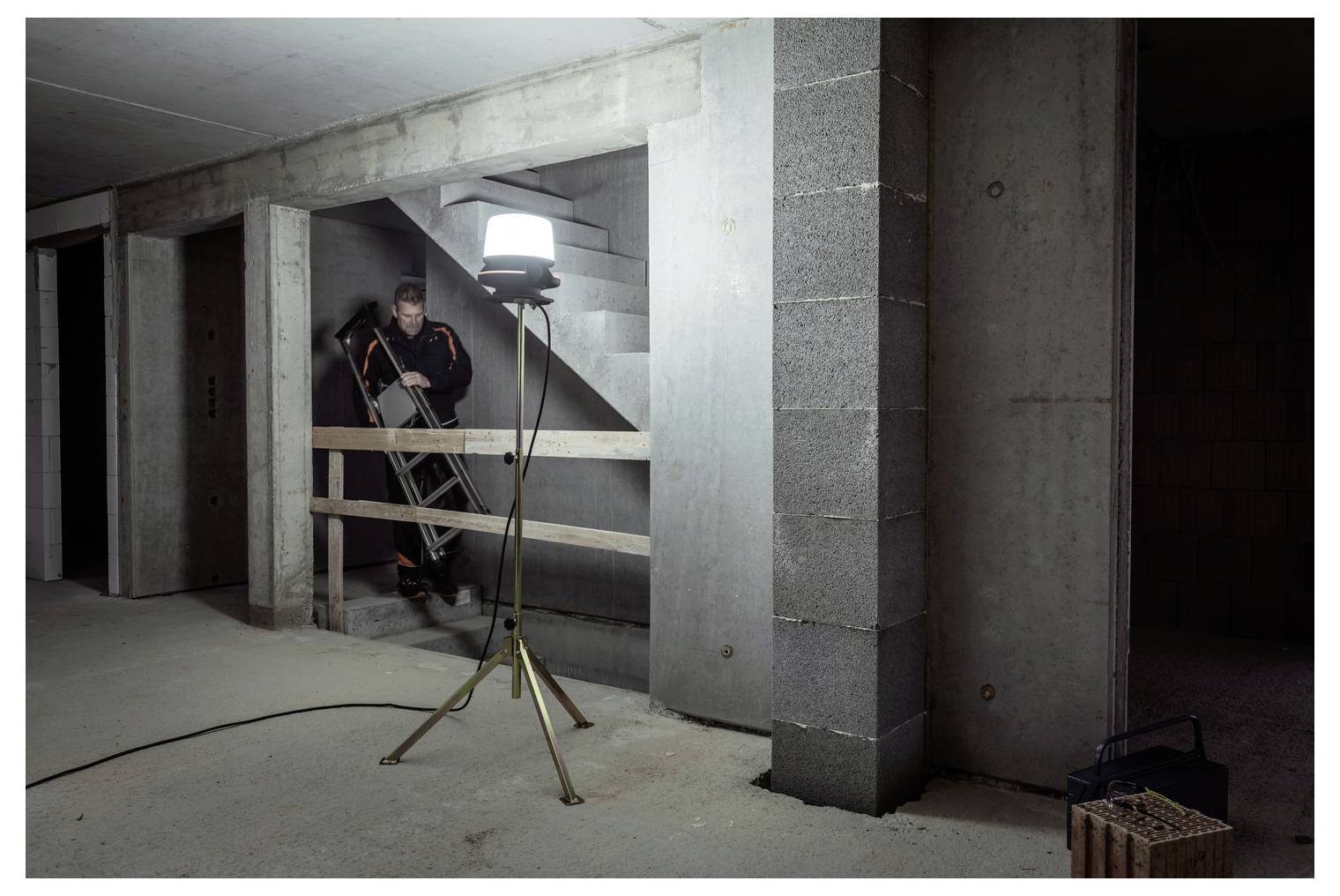 Bouwvakker op een ladder in een onafgewerkt betonnen souterrain, verlicht door een statief-werklamp in de buurt van de trap.