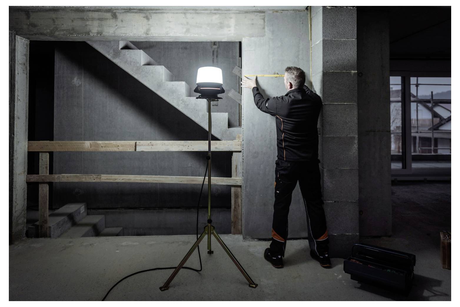 Een arbeider meet een betonnen muur in een onafgebouwd gebouw, verlicht door een statieflampje vlakbij een trap.
