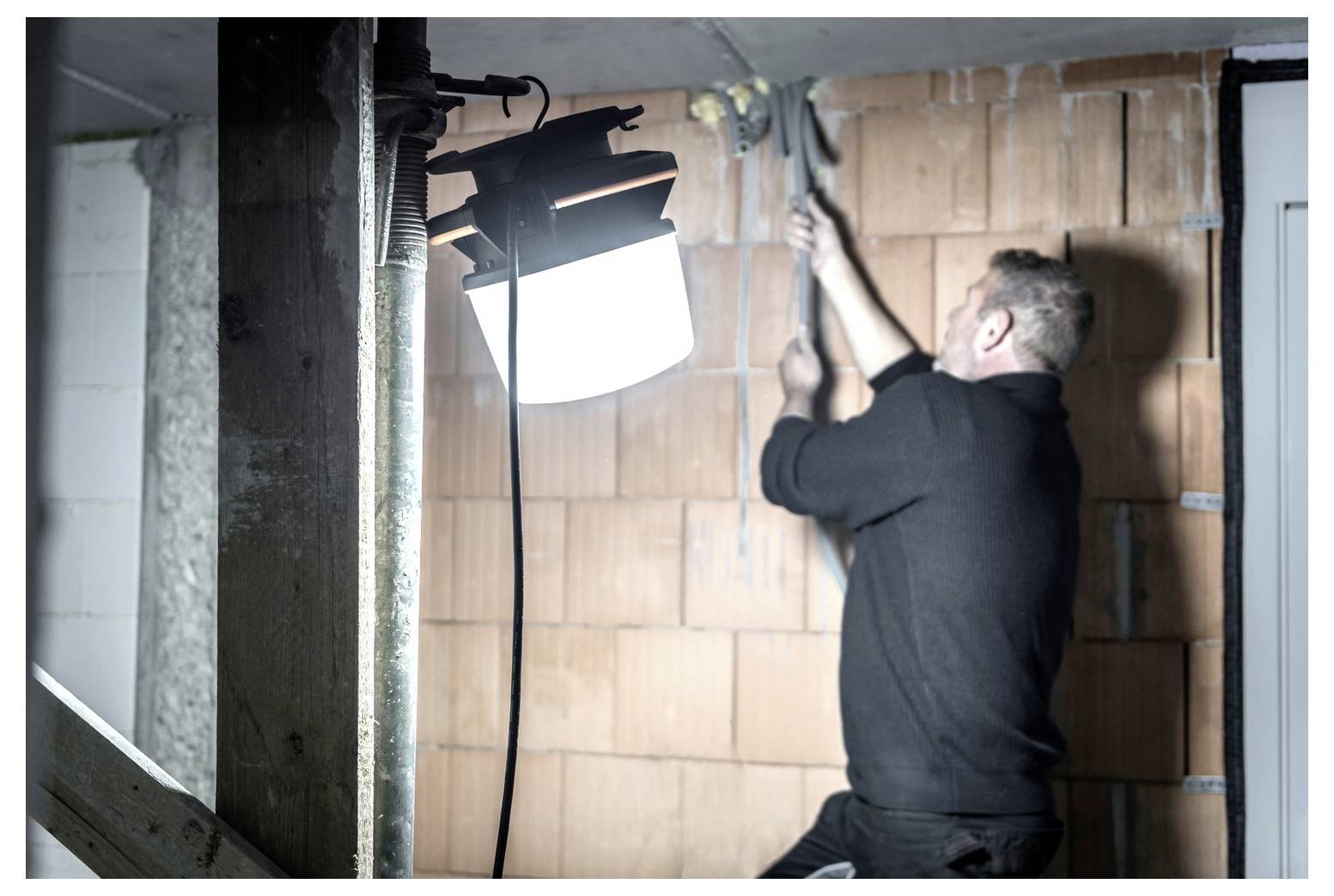 Een werknemer installeert of bevestigt een draad op een bakstenen muur terwijl een helder werklicht het gebied verlicht.