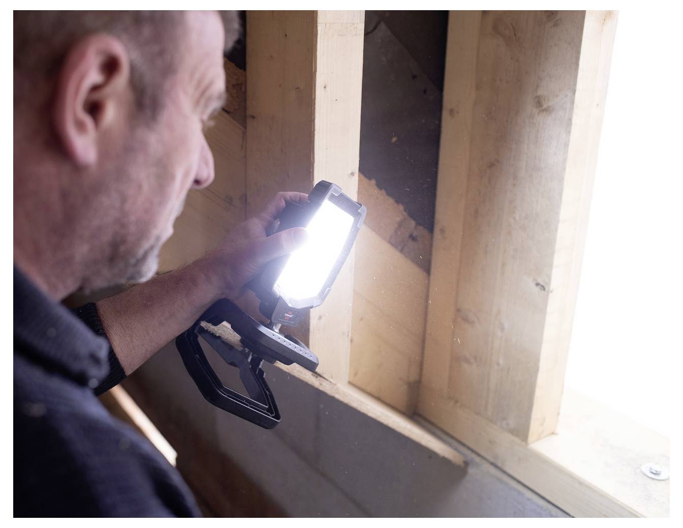 Man inspecteert een muurnisser met een handheld werklamp, controleert houten constructie in een gedeeltelijk ingebouwde binnenruimte.