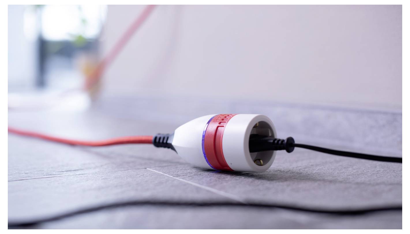 Close-up van een wit-rode elektrische stekker of connector liggend op een tegelvloer met rode en zwarte snoeren eraan bevestigd.
