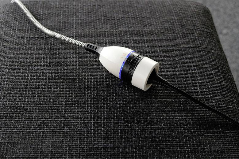 Een witte adapter met een zwart kabel op een grijze stoffen ondergrond. De stekkerverbinding heeft een blauwe lichtindicator.