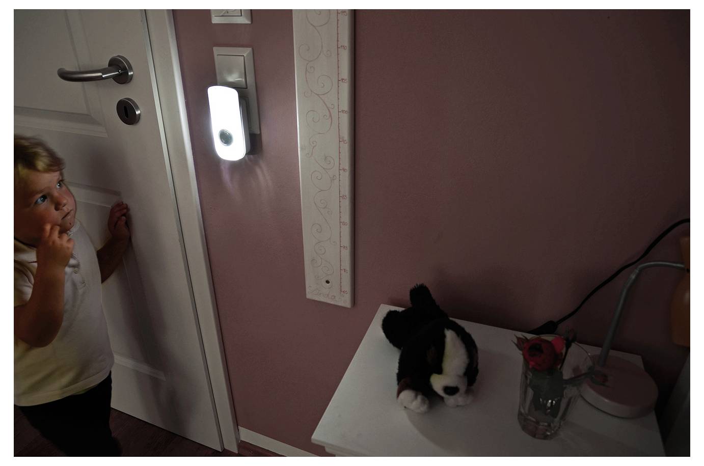 Klein kind gluurde achter een deur vandaan naar een klein zwart-wit knuffelkatje op een witte tafel naast een vaas en lamp in een roze kamer.