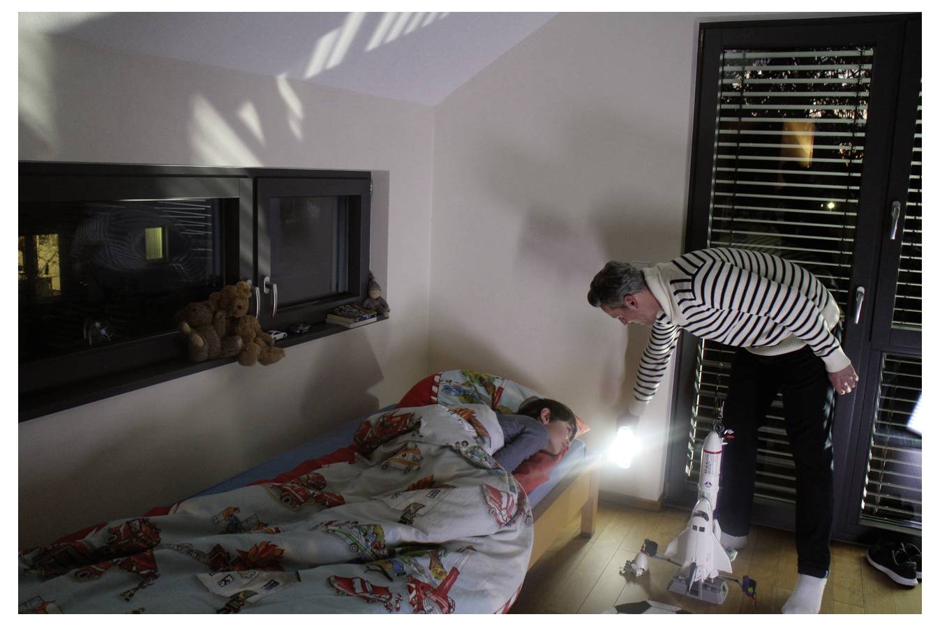 Een man gebruikt een zaklamp om 's nachts een slapend kind in bed te controleren, met een speelgoed raket ernaast dicht bij de deur.