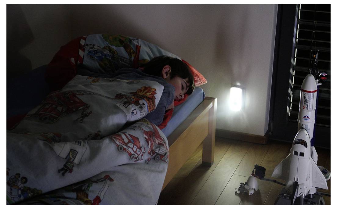 Kind dat slaapt in bed onder een deken terwijl een speelgoed ruimteshuttle en ander speelgoed op de grond naast het gloeiende nachtkastlampje staan.