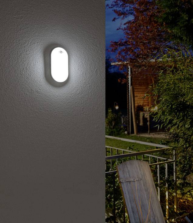 Een ovale wandlamp schijnt in het donker. Op de achtergrond is een verlichte tuin met terras en bomen zichtbaar.