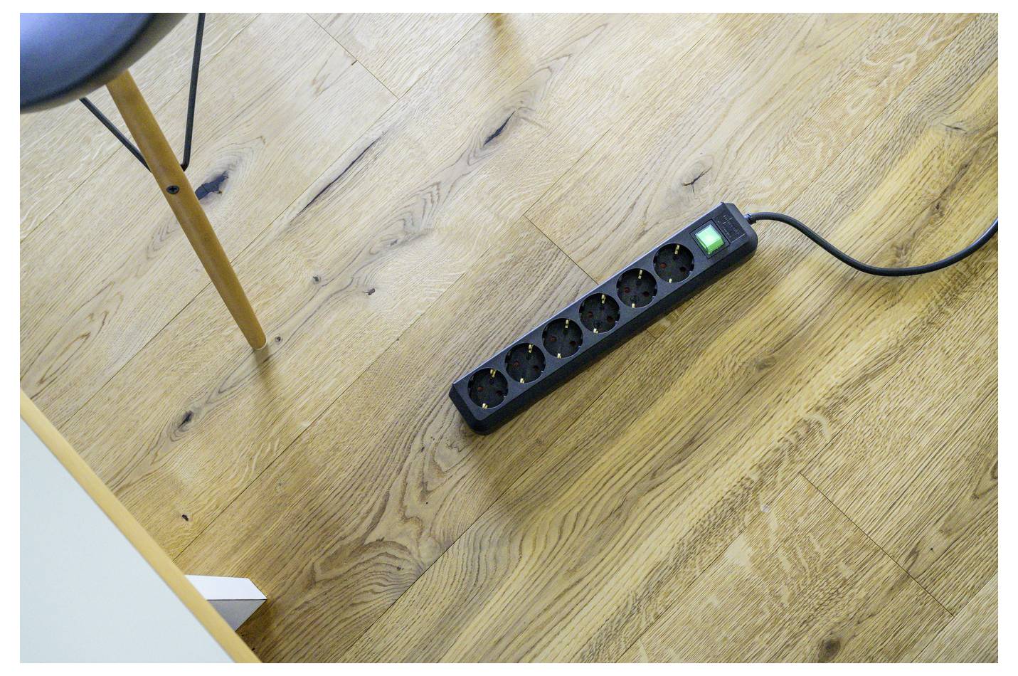 Zwart stroomverdeelblok met zes stopcontacten en een gloeiende groene schakelaar ligt op een houten vloer naast een stoelpoot.