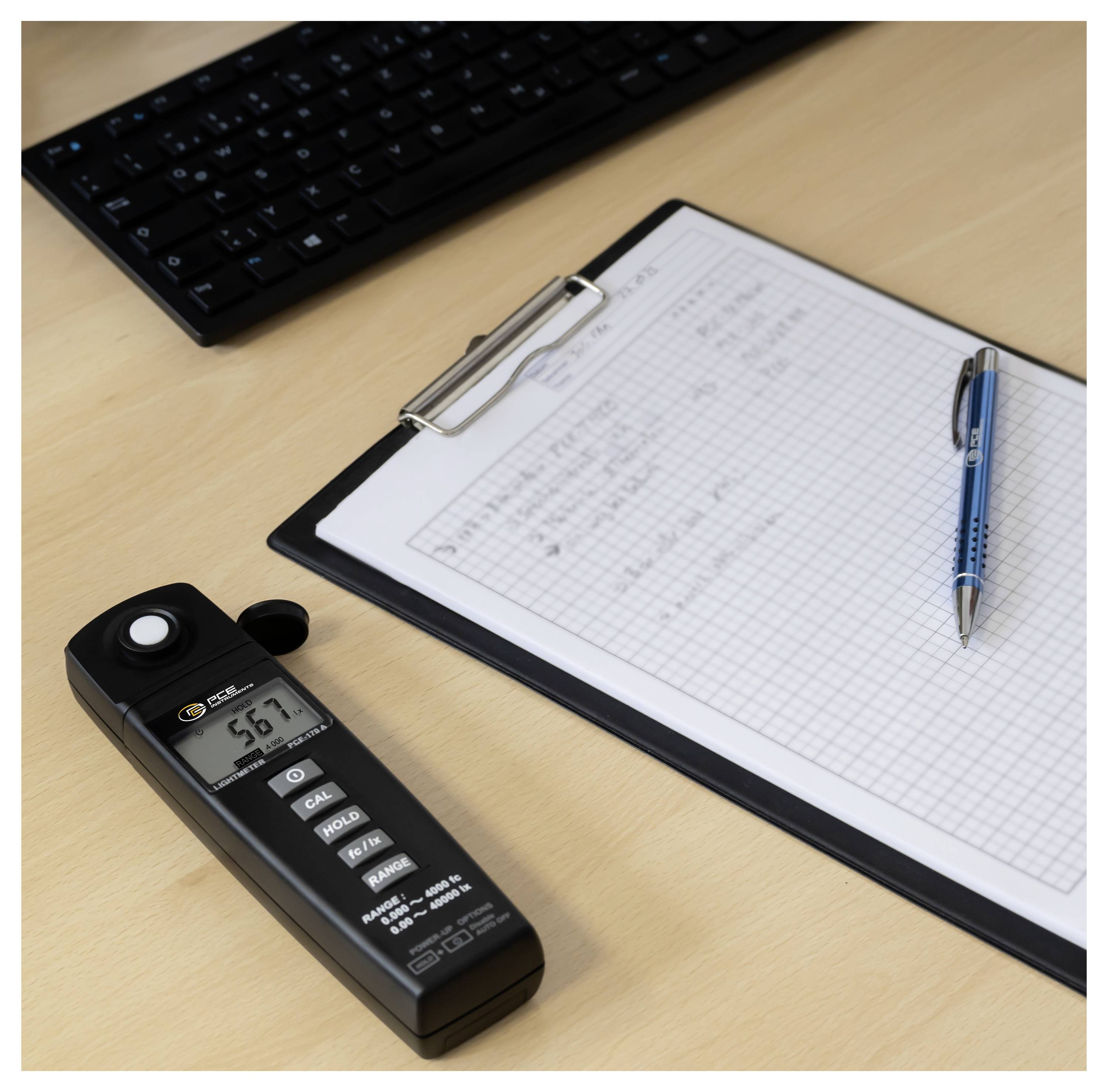 Luxmeter lezing 56,7 op een bureau naast een klembord met millimeterpapier, een pen en een toetsenbord.