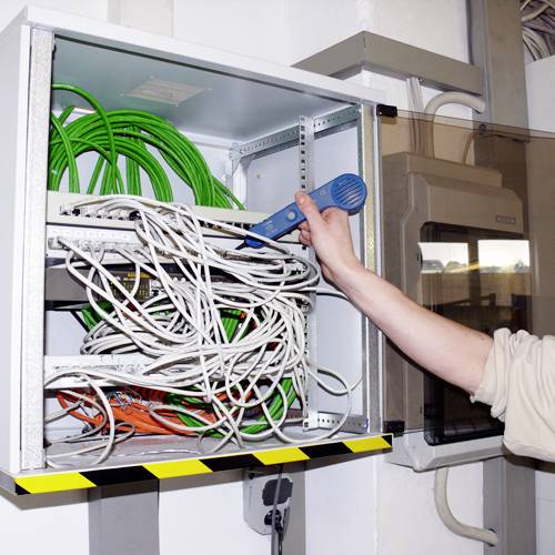 Een persoon gebruikt een blauw gereedschap om een kabel te testen in een open schakelkast met veel groene en witte kabels.