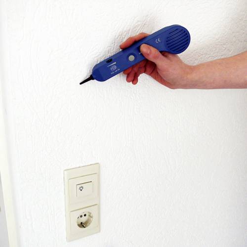 Een hand houdt een blauw apparaat vast tegen een witte muur boven een stopcontact.