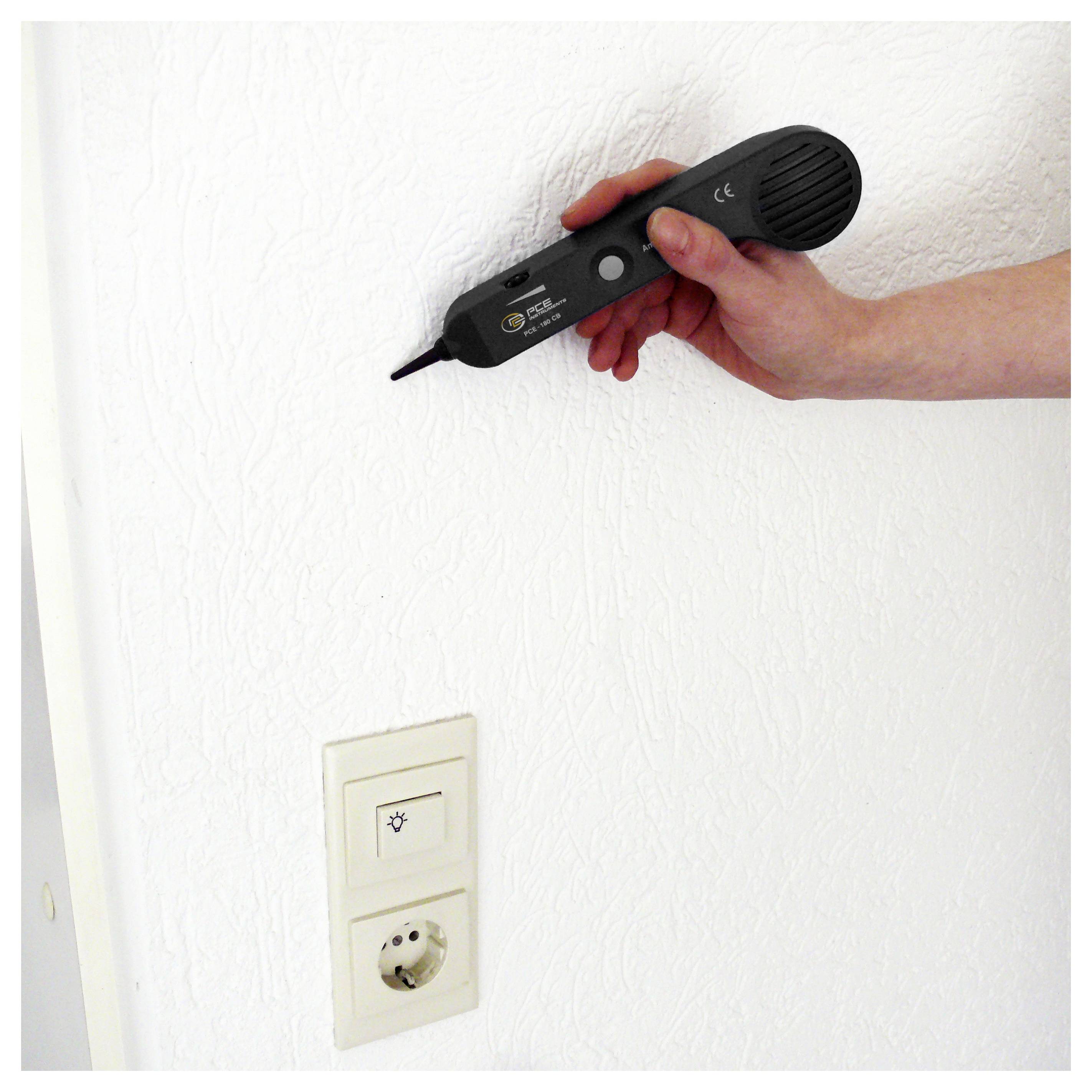 Hand die een spanningsproefer vasthoudt dicht bij een muur boven een lichtschakelaar en stopcontact, waarbij de muur wordt gecontroleerd op onder spanning staande bedrading.