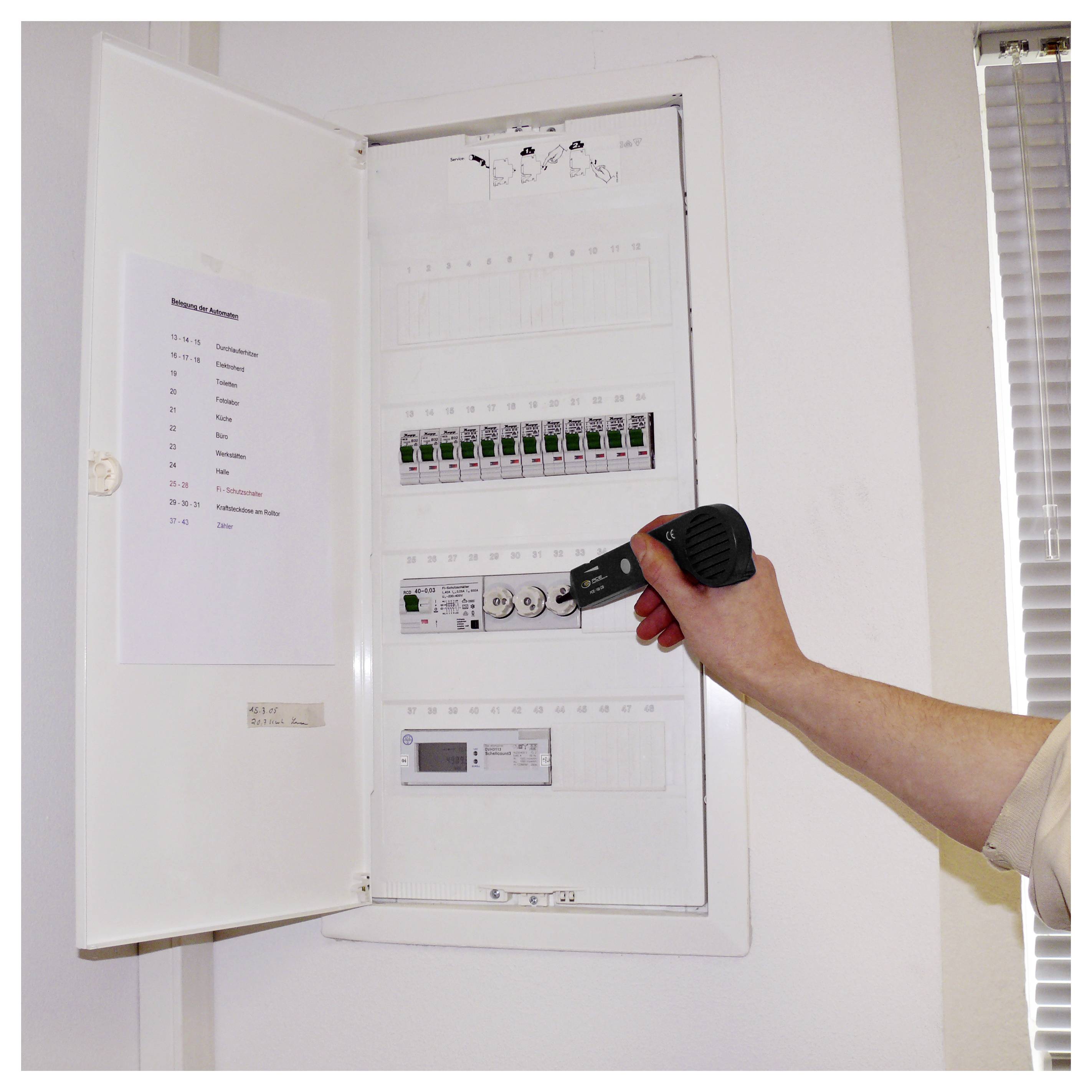 Hand die een zaklamp vasthoudt en een open elektrisch zekeringkastje met gelabelde schakelaarsen in een witte wandkast inspecteert.