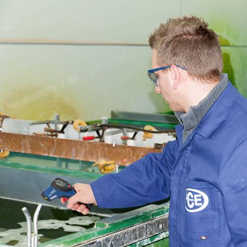 Een man in een blauwe laboratoriumjas meet de temperatuur van een vloeistof in een industrieel bad met een thermometer.