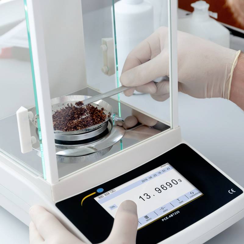 Een persoon weegt koffiebonen op een precisieweegschaal. De aanduiding toont 13,9680 gram aan.