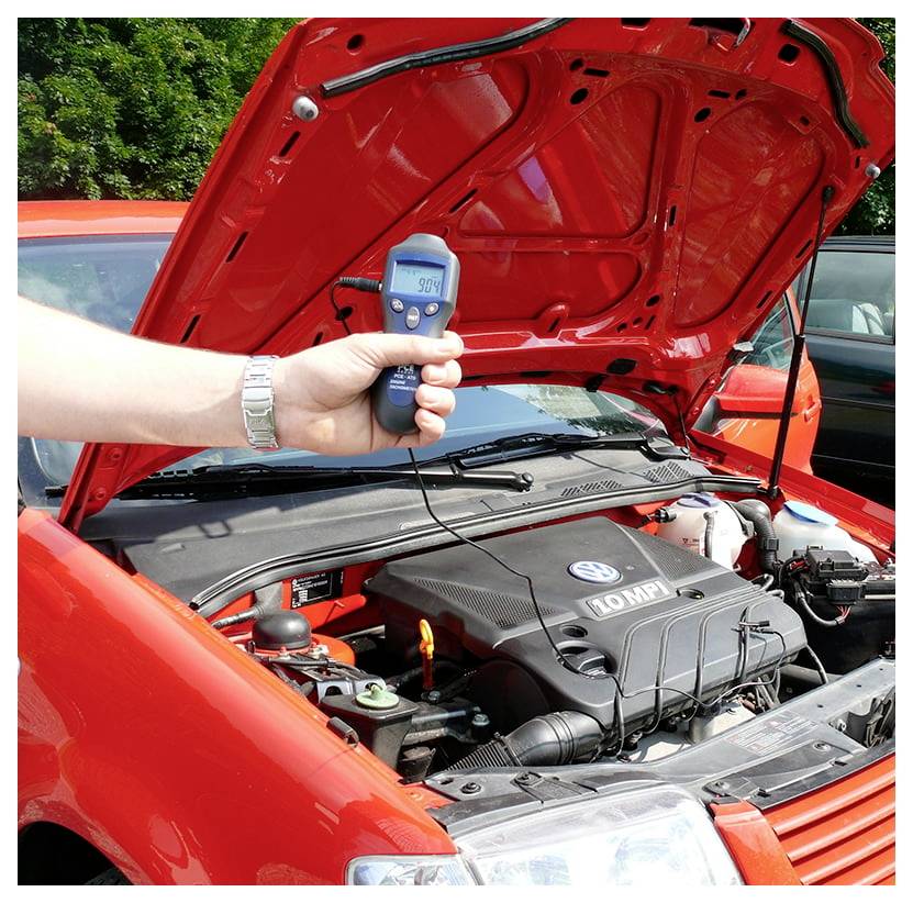 Draagbare infraroodthermometer gericht op de open motorkap van een rode auto, met een temperatuurweergave terwijl de motorhitte wordt gecontroleerd.
