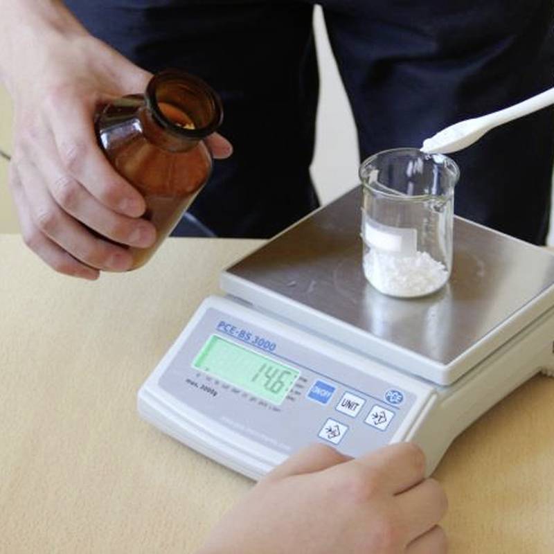Een glazen potje met wit poeder wordt gewogen op een elektronische weegschaal. Een hand houdt een bruine flesje vast. Gewichtsaanduiding: 14,6 g.