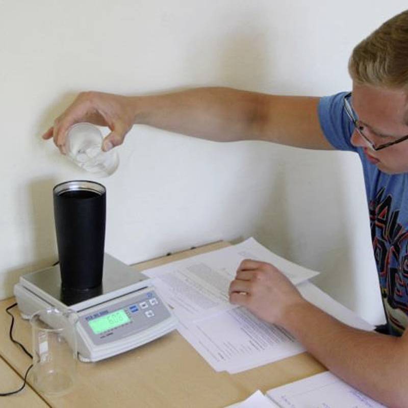 Een persoon giet water uit een maatbeker in een zwarte container op een digitale weegschaal die op een tafel staat.