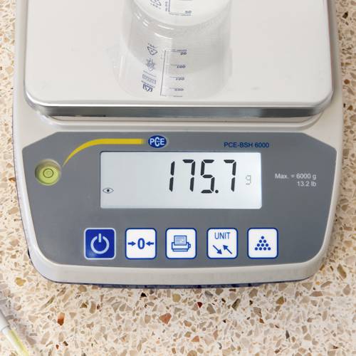 Elektronische weegschaal geeft 175,7 g aan. Heldere plastic schaal met vloeistof bovenop. Weegschaal op beige gevlekte ondergrond.